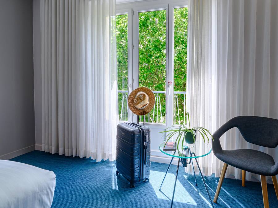 Een hotelkamer met een groot raam, een blauw tapijt, een zwarte koffer, een ronde hoed op een standaard, een glazen tafel met een plant en een moderne stoel.