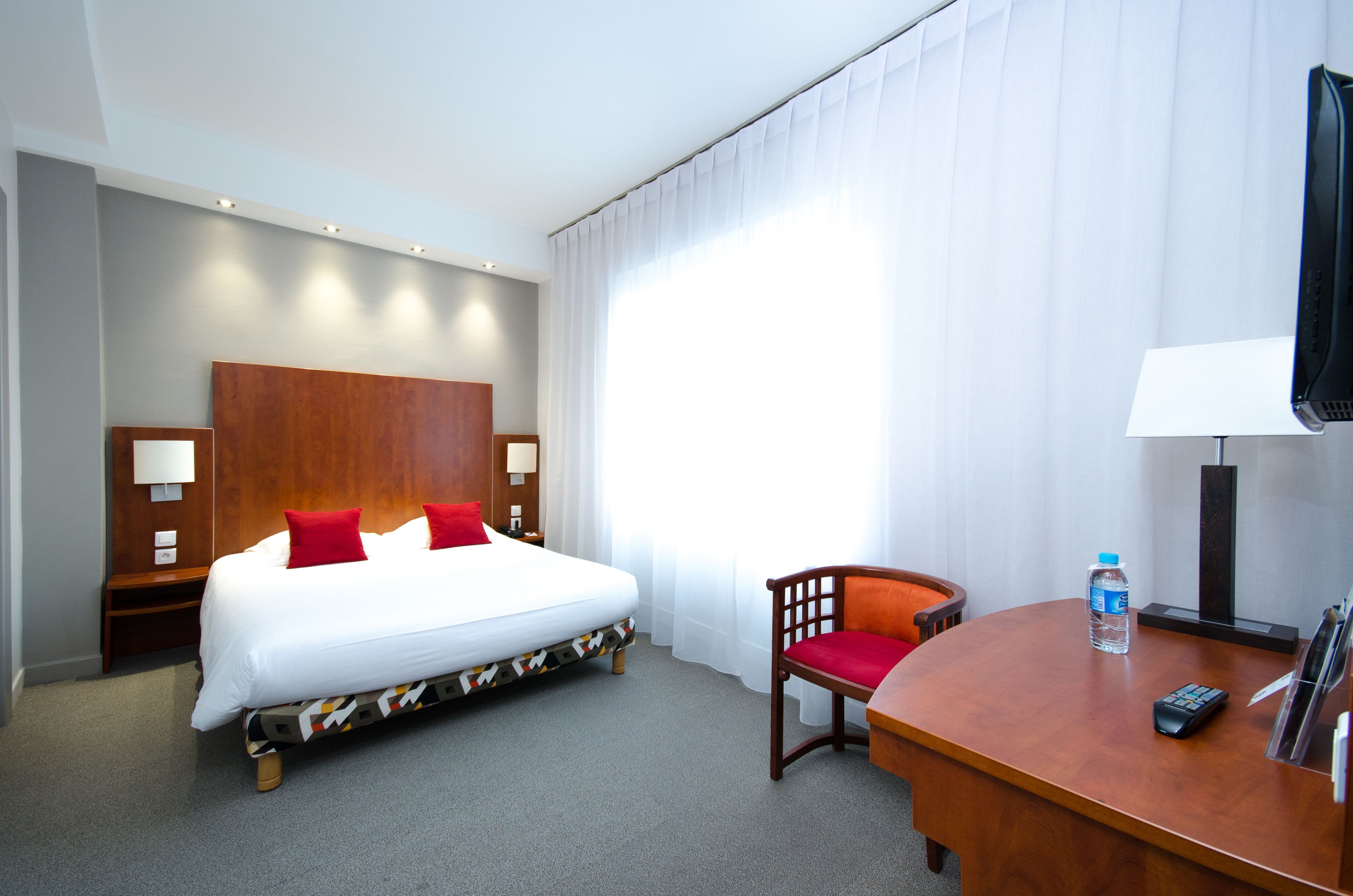 A hotel room with a bed, a desk, a chair, a TV, and a lamp.