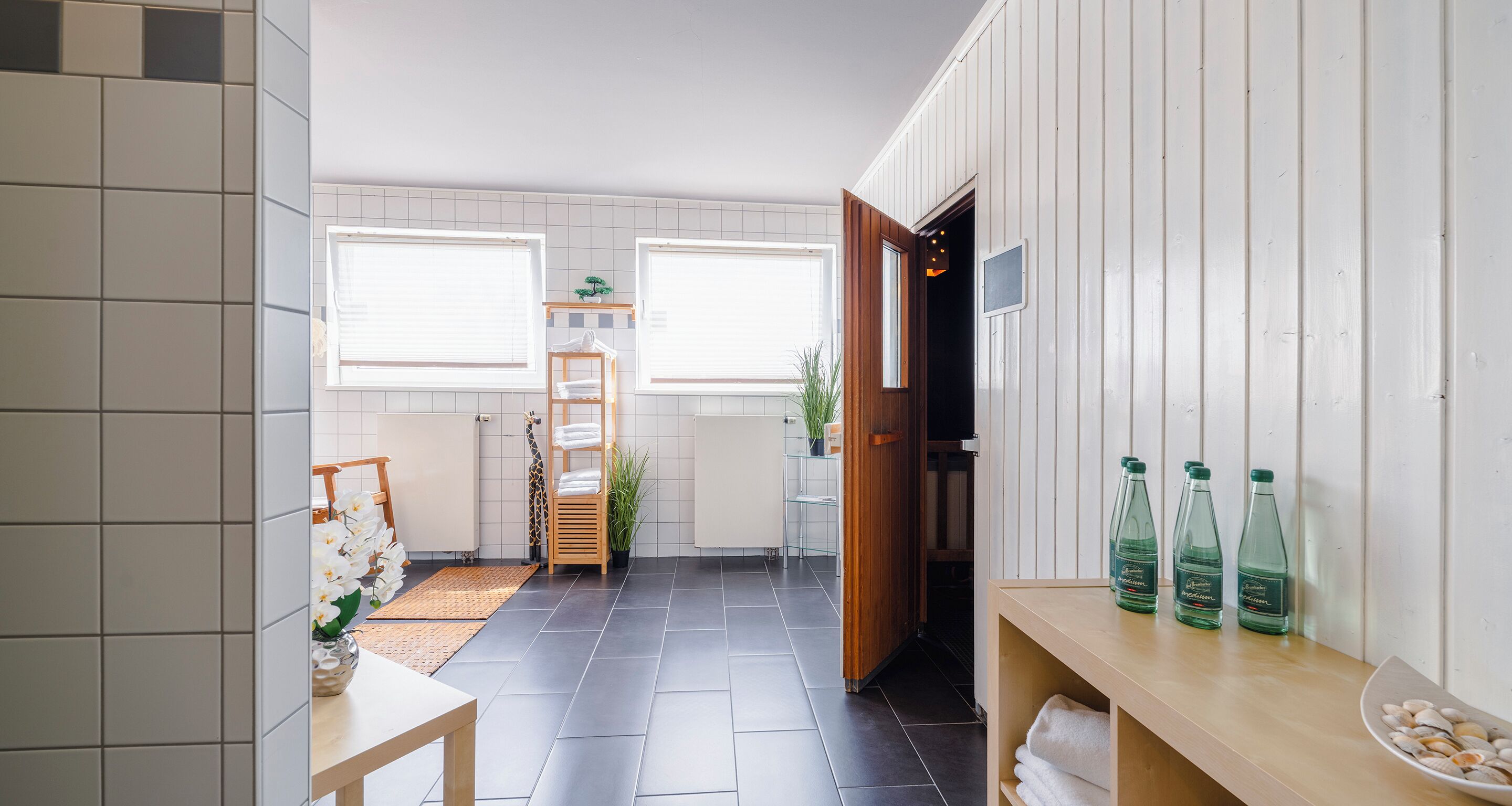 A modern bathroom with a tiled floor, a wooden bench, a shelf with bottles, and a towel rack.