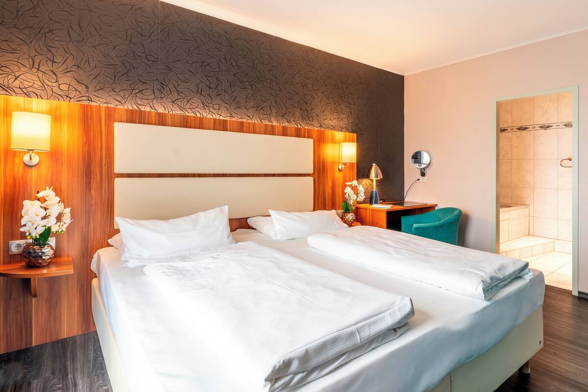 Una habitación de hotel con dos camas, un cabecero de madera, una mesita de noche con lámpara, un espejo y un baño con ducha.