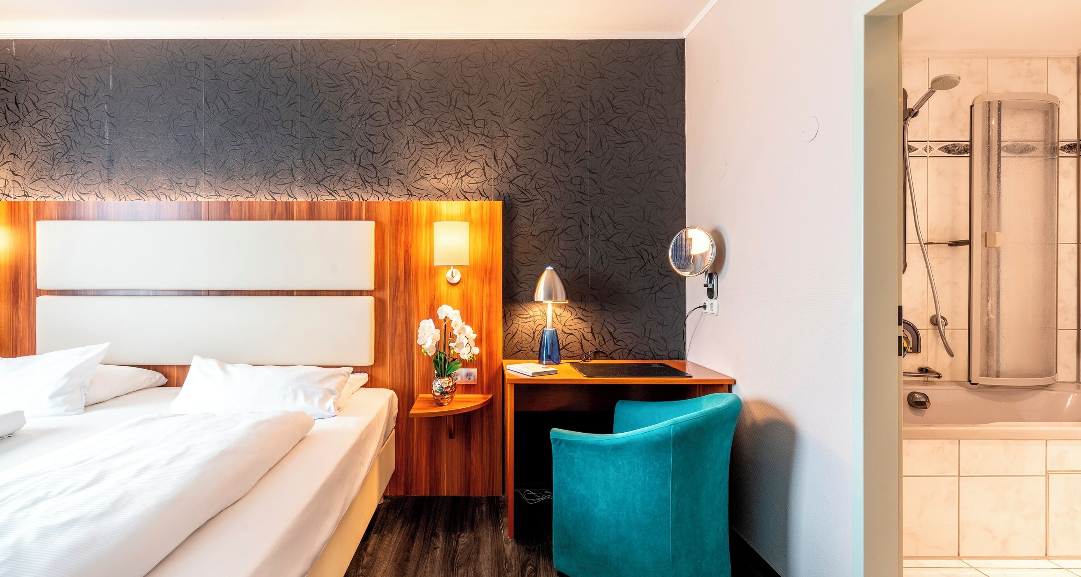 A hotel room with a bed, a desk with a lamp, a chair, and a shower area.