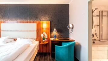 A hotel room with a bed, a desk with a lamp, a chair, and a shower area.