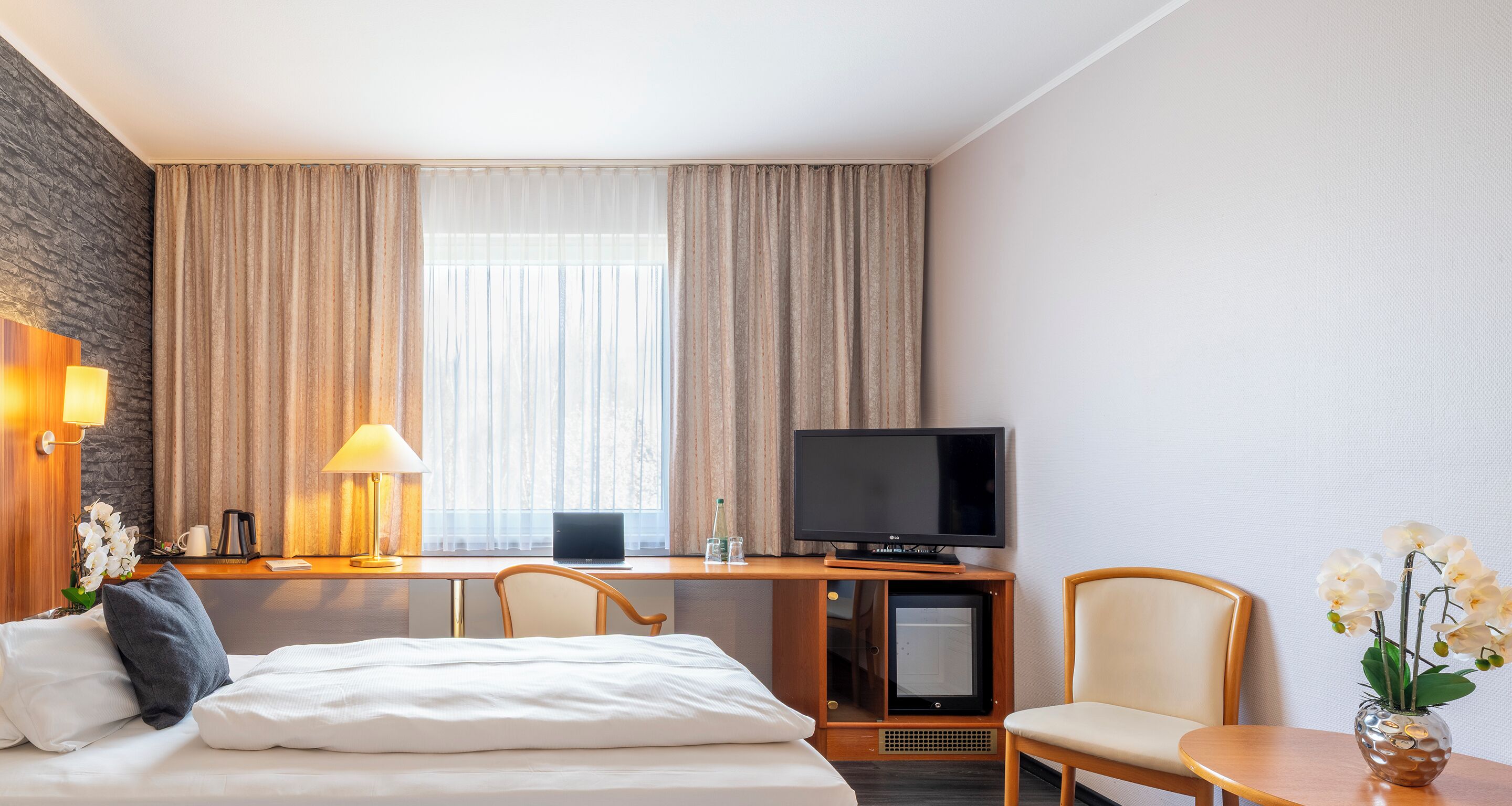 Een hotelkamer met een bed, een bureau met een laptop, een televisie, een stoel en een tafeltje met een bloemenvaas.