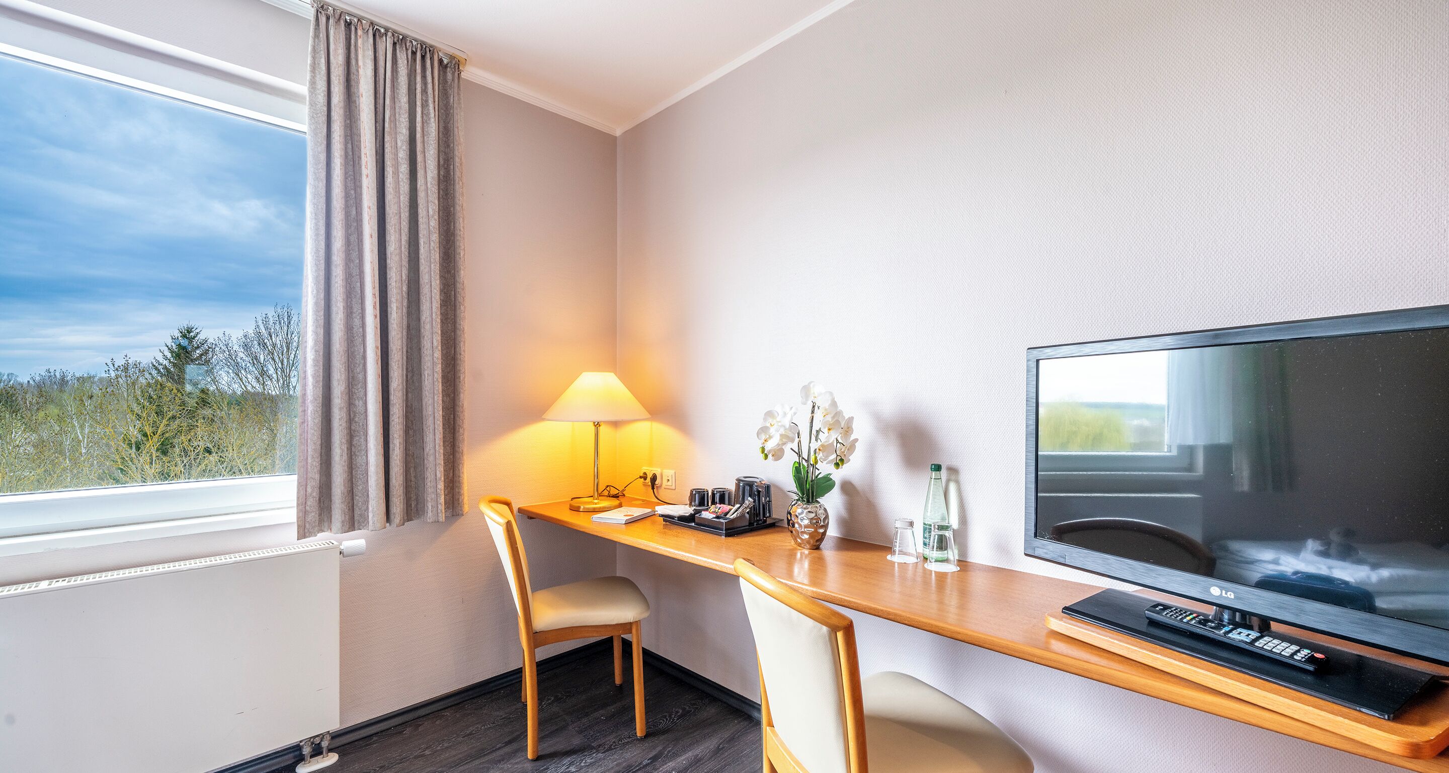 A hotel room with a desk, two chairs, a TV, a lamp, a vase with flowers, and a window with curtains.
