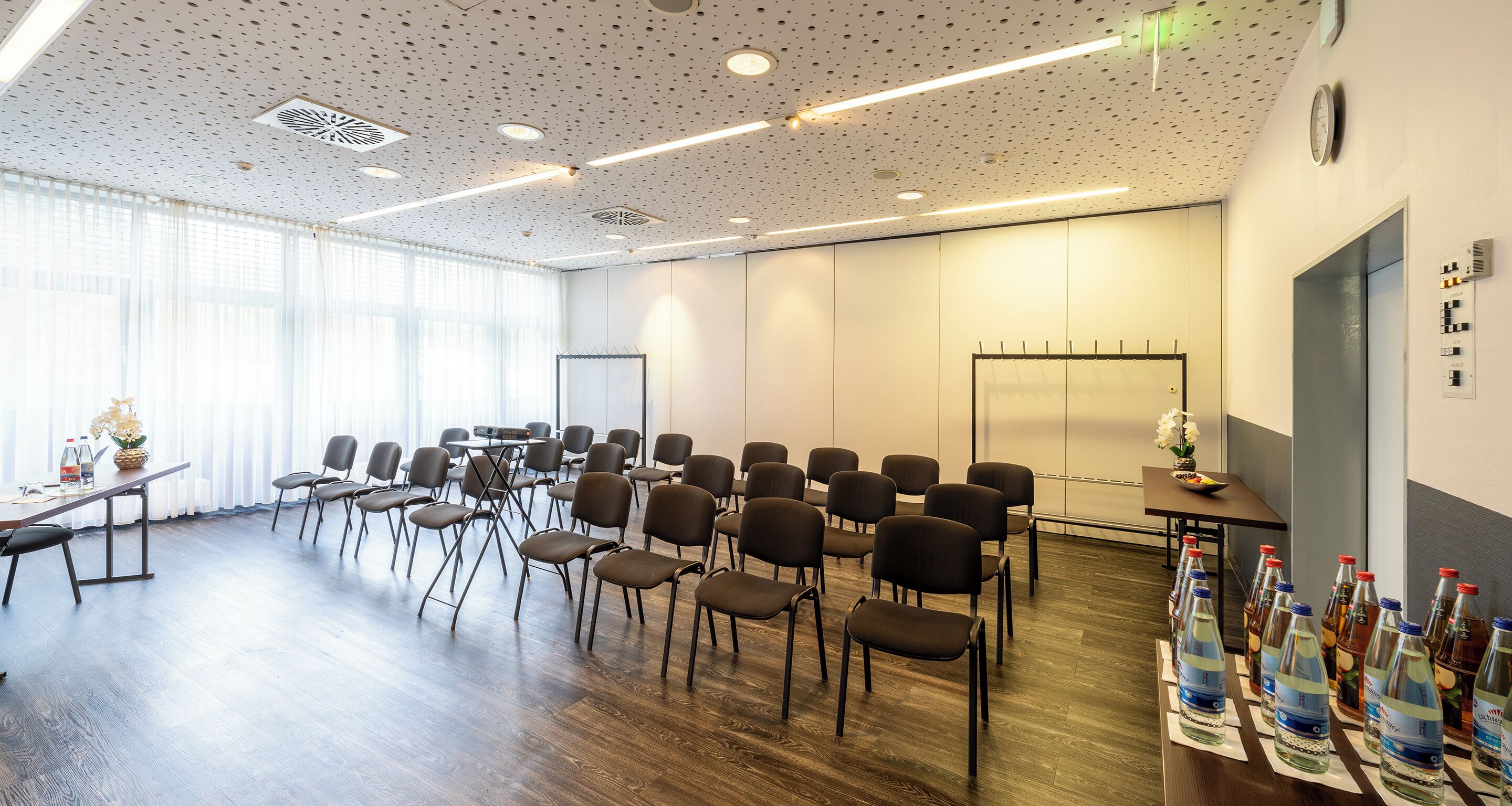 Un salón de conferencias con filas de sillas, una mesa con botellas de agua y un reloj en la pared.