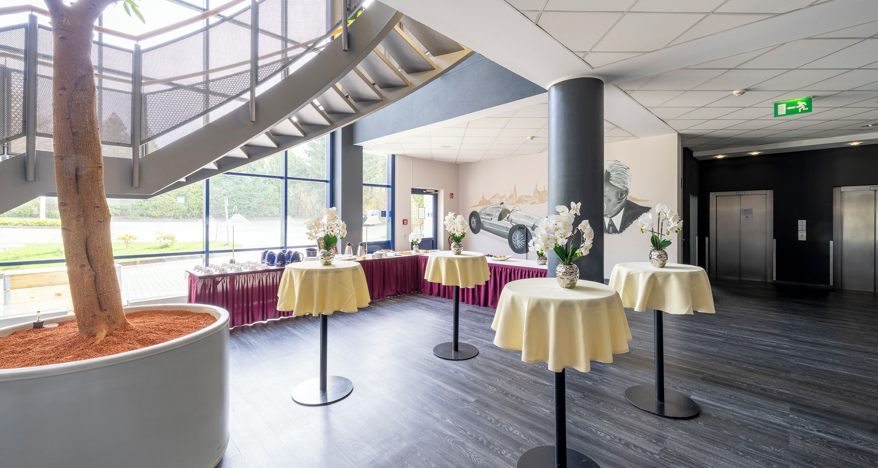 Een moderne lobby met een grote potplant, meerdere tafels met gele tafelkleden, en een trap.