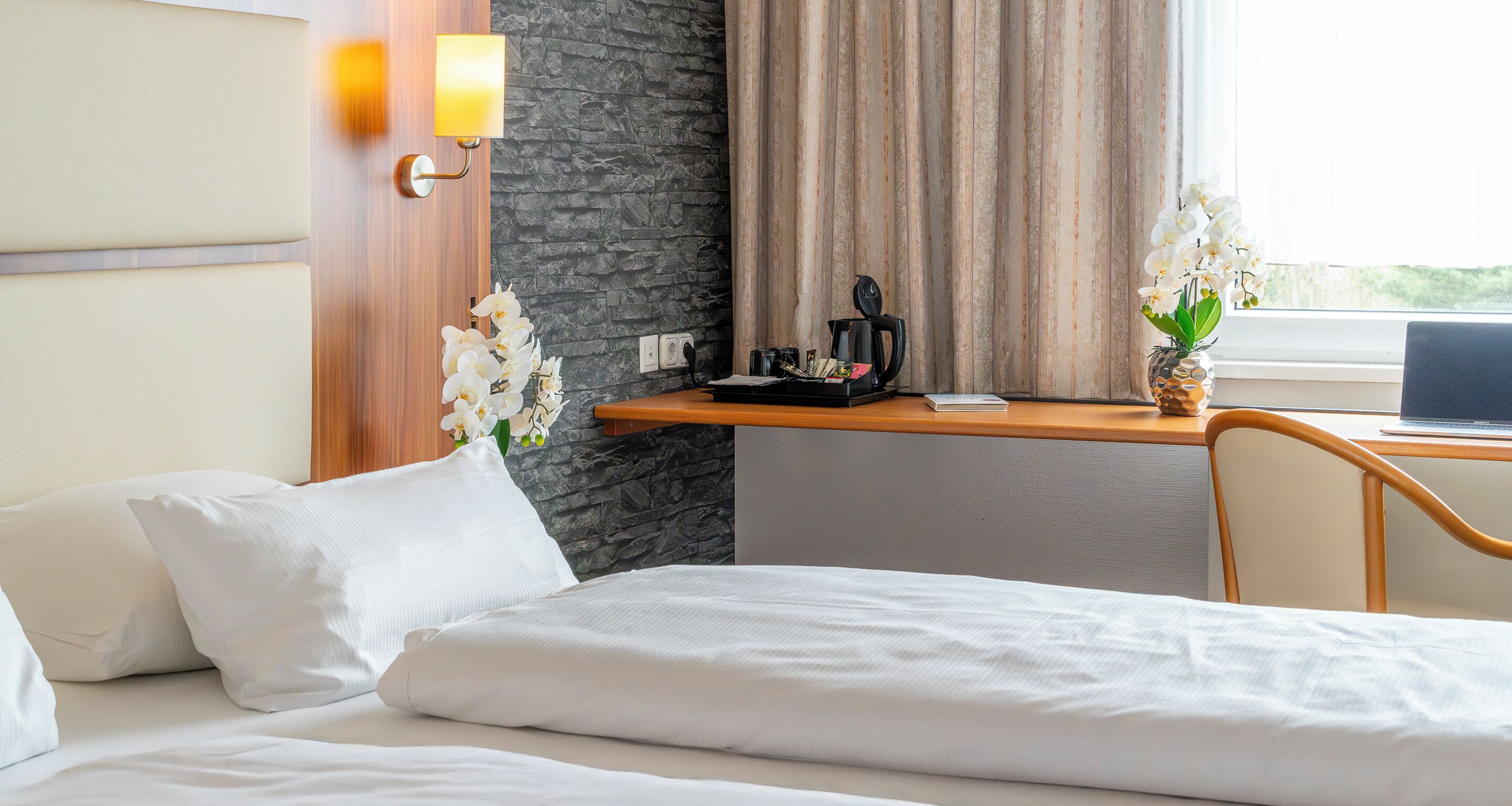Ein Hotelzimmer mit einem Bett, einem Schreibtisch mit einem Laptop, einer Kaffeemaschine und weißen Orchideen.