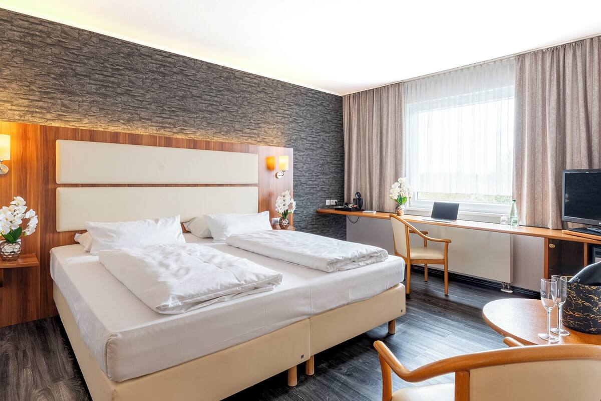 Habitación de hotel con una cama, un escritorio con computadora, televisor y ventana con cortinas.