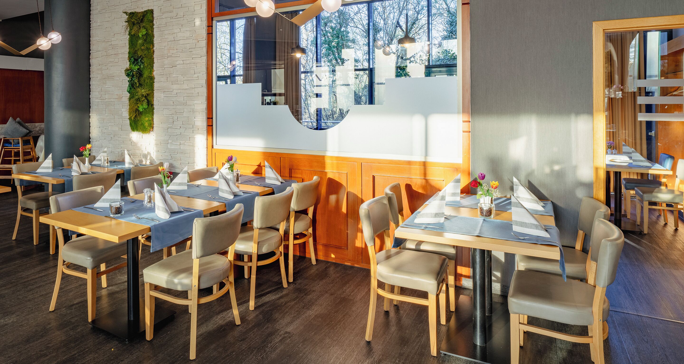 Un interior de restaurante moderno con mesas y sillas de madera, un jardín vertical verde y grandes ventanales que ofrecen vistas al exterior.