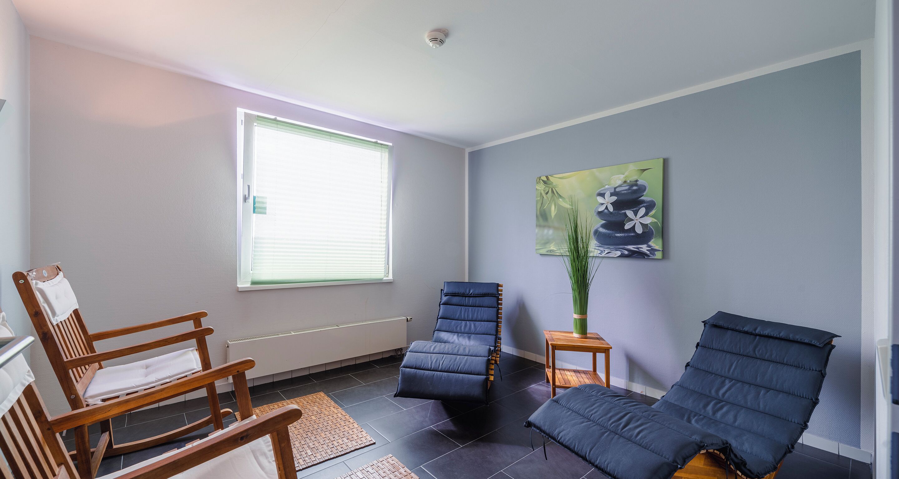 Una habitación con una mecedora de madera, un sillón azul acolchado y un sillón reclinable azul. Hay una ventana con persianas, un radiador y un cuadro en la pared.