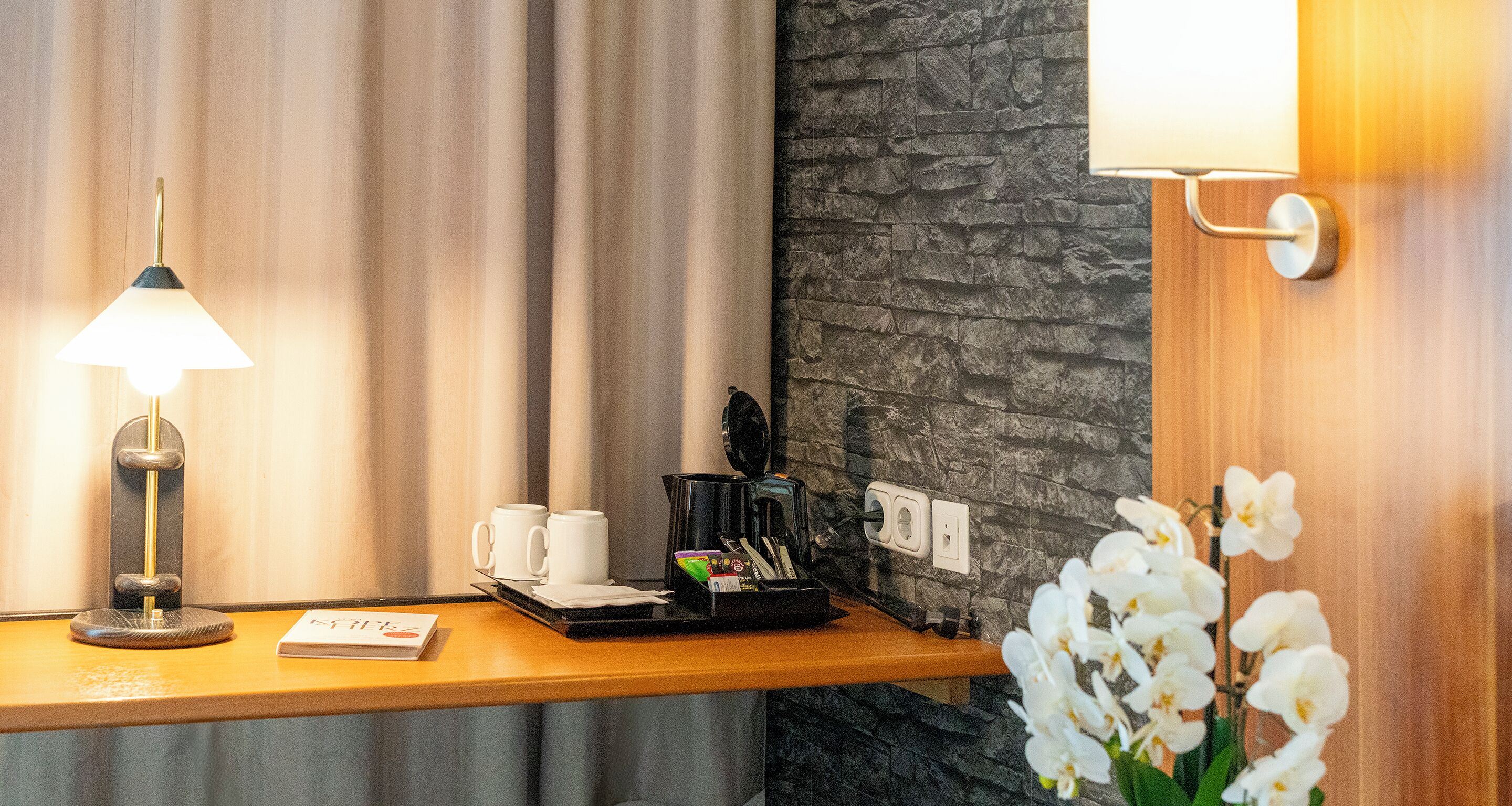 A hotel room with a wooden desk, a lamp, two white cups, a coffee maker, and a flower arrangement.