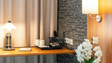 A hotel room with a wooden desk, a lamp, two white cups, a coffee maker, and a flower arrangement.