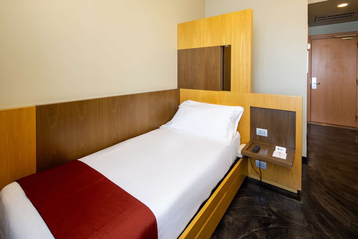 A single bed with a wooden headboard, a bedside table with a phone and a notepad, and a red throw blanket.
