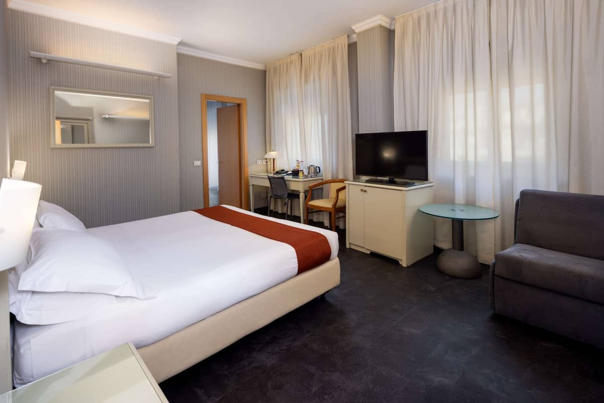 A hotel room with a large bed, a desk with a TV, a chair, and a small table.