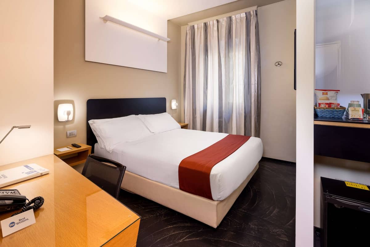 A hotel room with a bed, bedside table, lamp, and a desk with a telephone and a name tag.