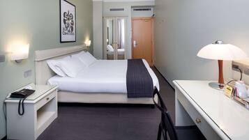 Quarto de hotel com cama, mesa de cabeceira com telefone, escrivaninha com abajur e espelho.