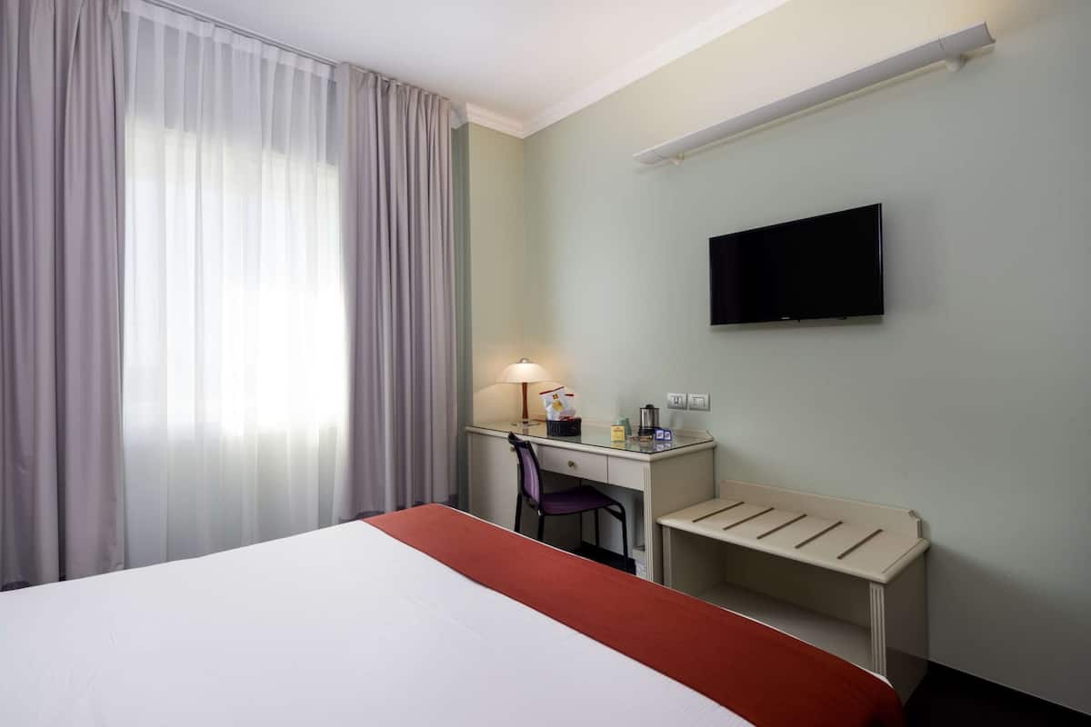 A hotel room with a bed, a desk, a chair, a TV, and a window with curtains.