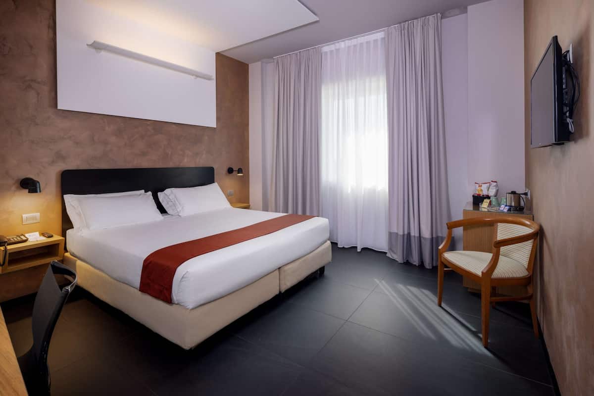 A hotel room with a large bed, a desk, a chair, a TV, and a window with curtains.