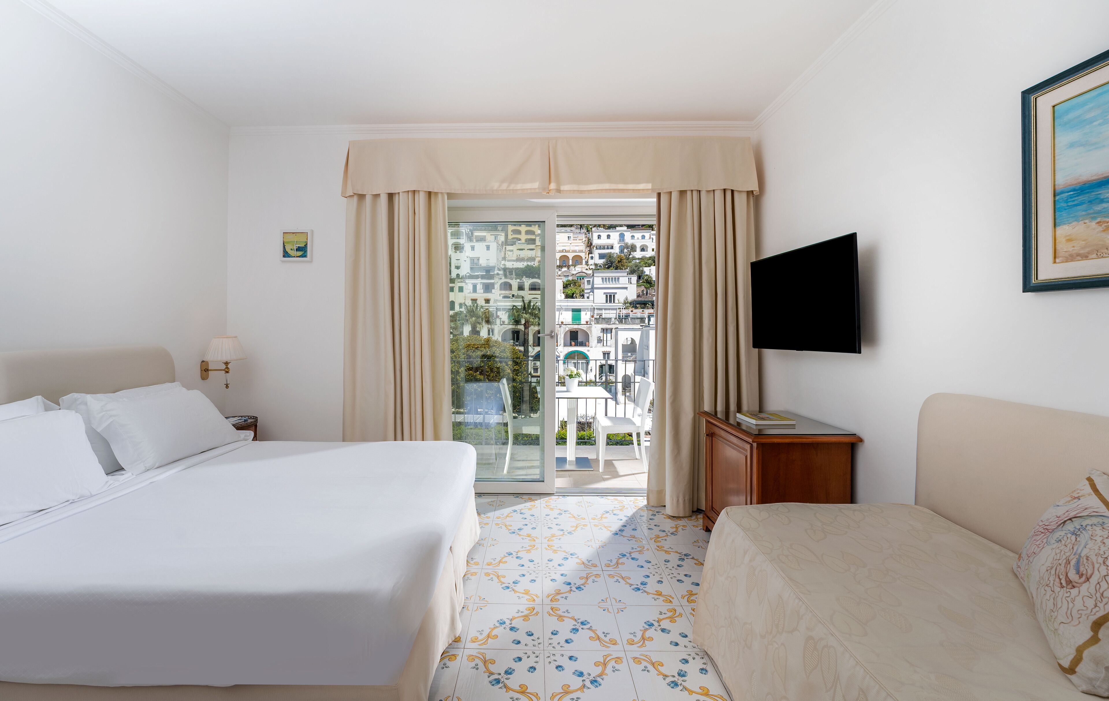 A hotel room with a bed, a desk, a TV, and a balcony with a view of buildings.