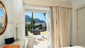 A hotel room with a balcony, a bed, a desk with a lamp, a book, and a view of the city and mountains.