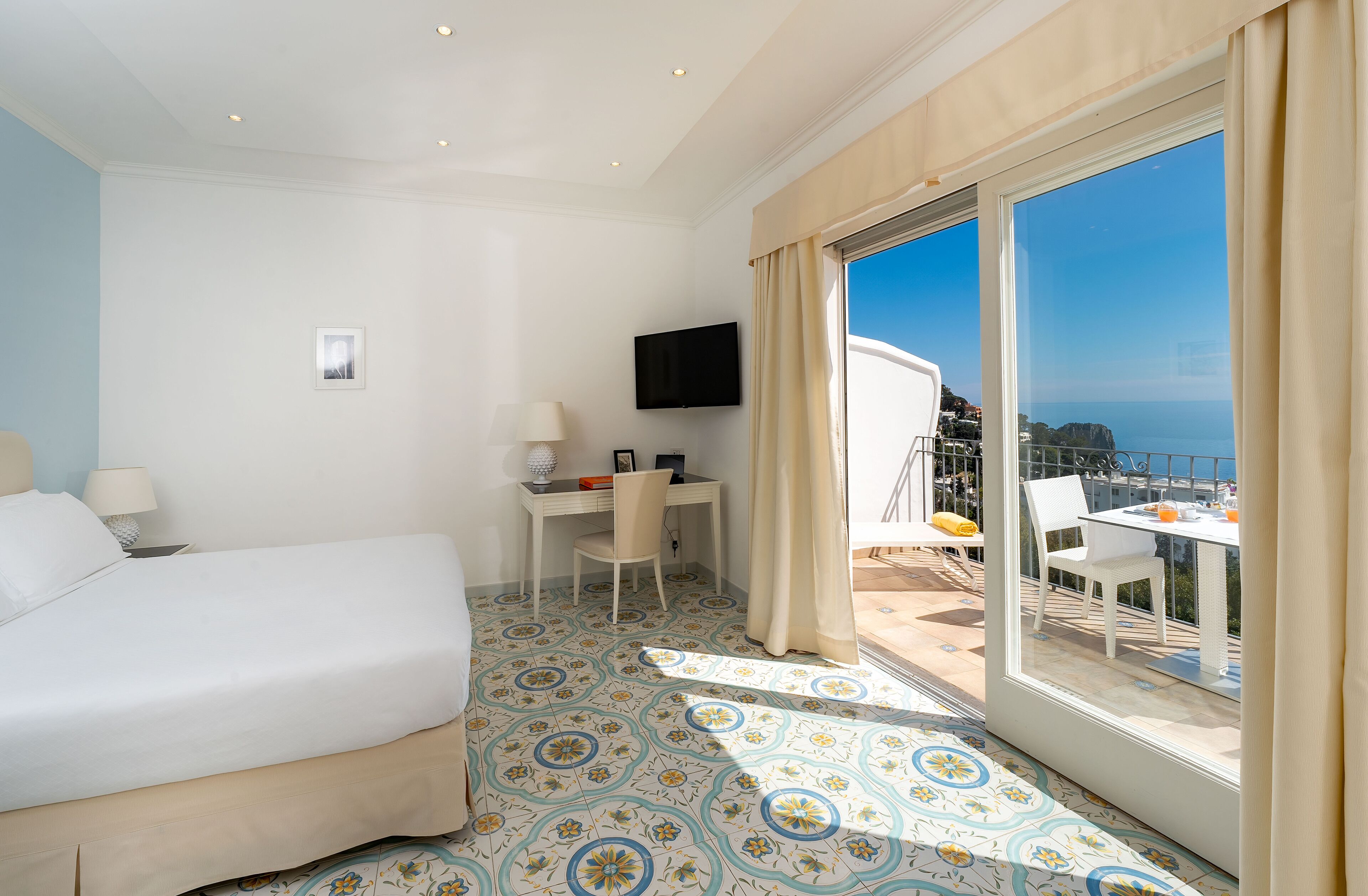 A hotel room with a bed, desk, TV, and a balcony with a table and chairs.