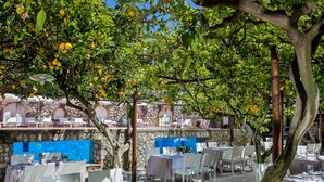 An outdoor dining area with white tables and chairs, surrounded by trees and a stone wall.