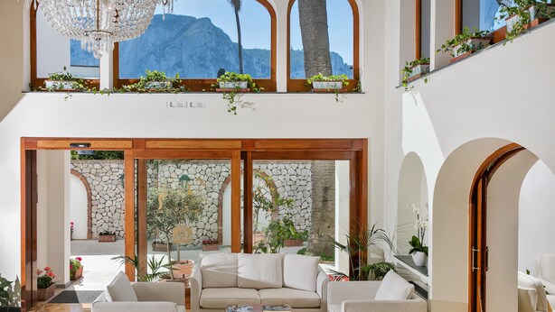 A spacious living area with a beige sofa, a coffee table, and potted plants. There are large windows with plants on the sill and a view of mountains.