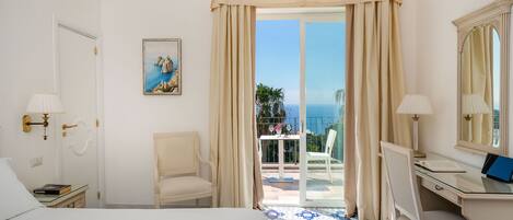A hotel room with a bed, a desk with a laptop, a chair, a balcony with a view of the sea, and a painting on the wall.