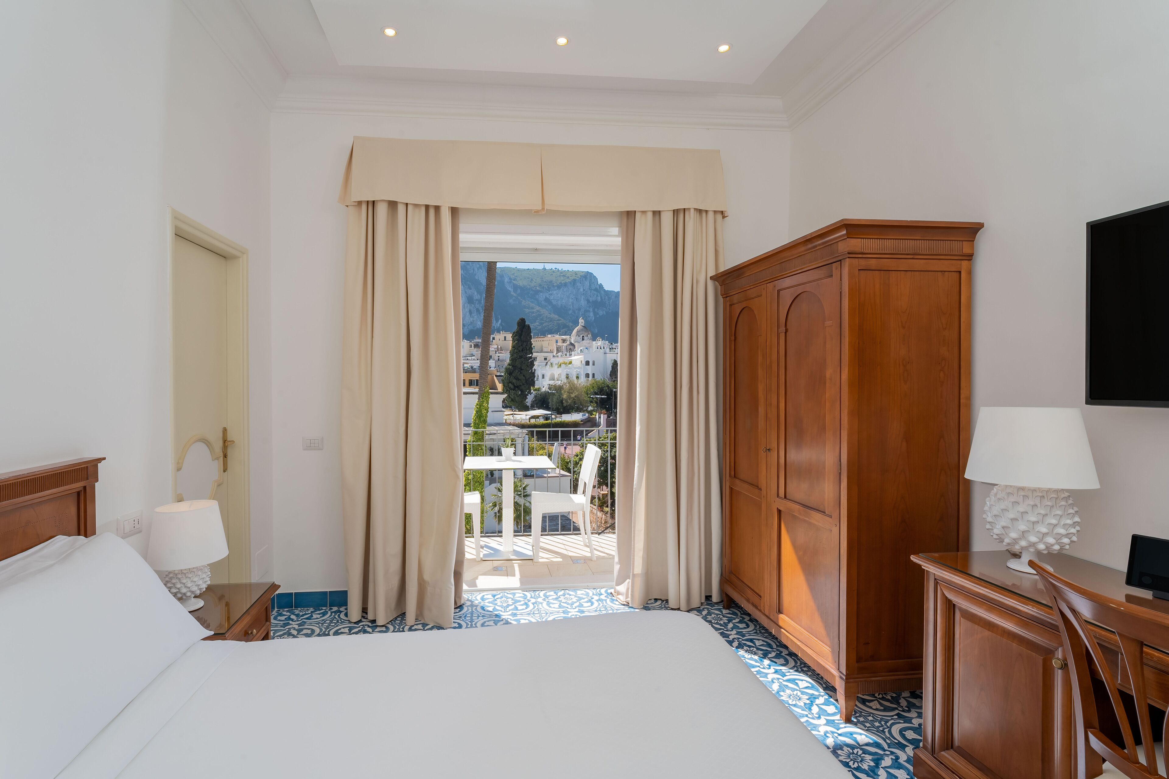 A hotel room with a bed, a wooden wardrobe, a desk with a lamp, and a view of a balcony with outdoor furniture.