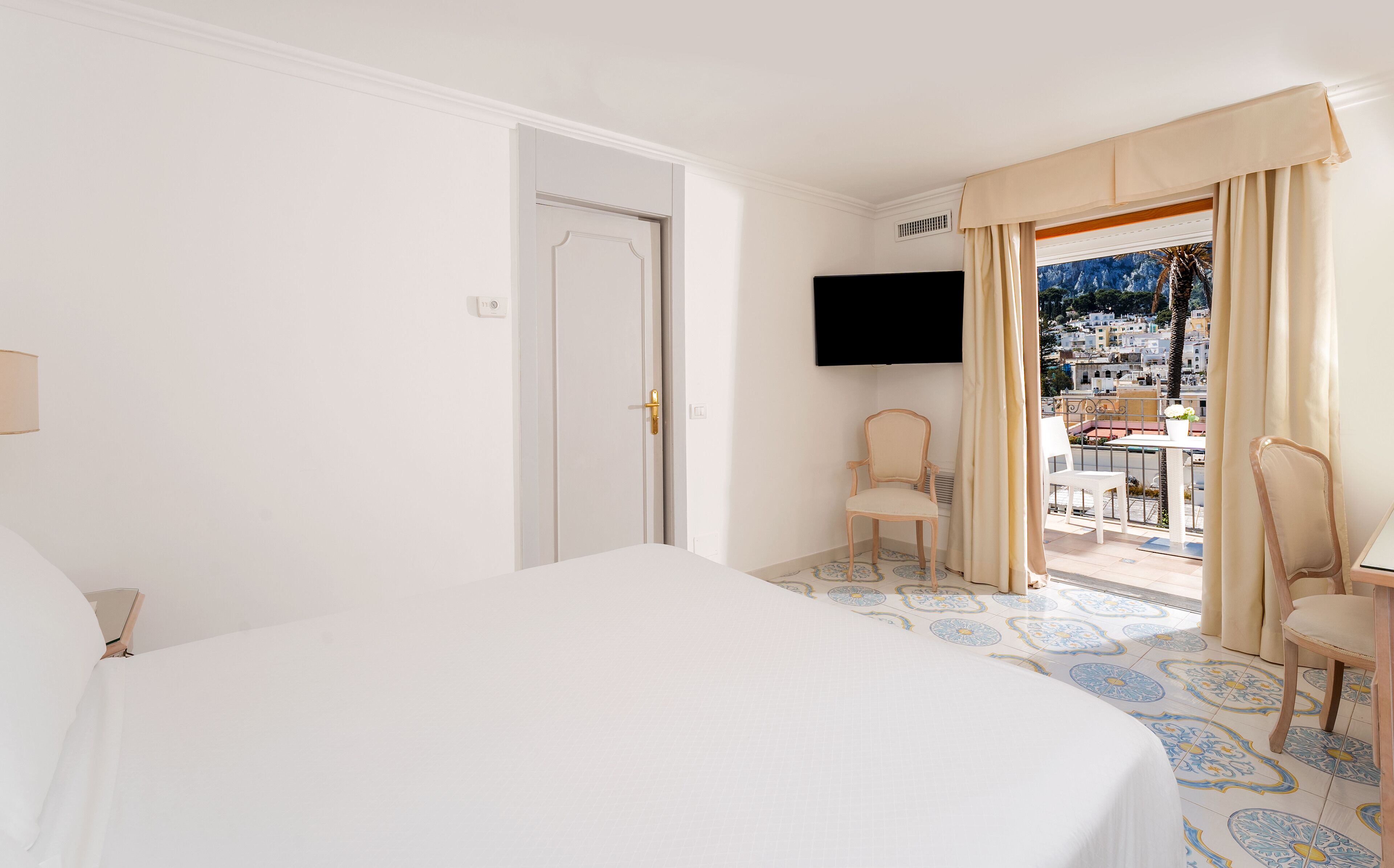 Ein Hotelzimmer mit Bett, einem an der Wand befestigten Fernseher und einem Balkon mit Blick auf die Stadt.