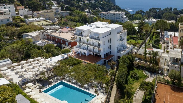 An aerial view of a coastal hotel with multiple floors, a swimming pool, and a tennis court.