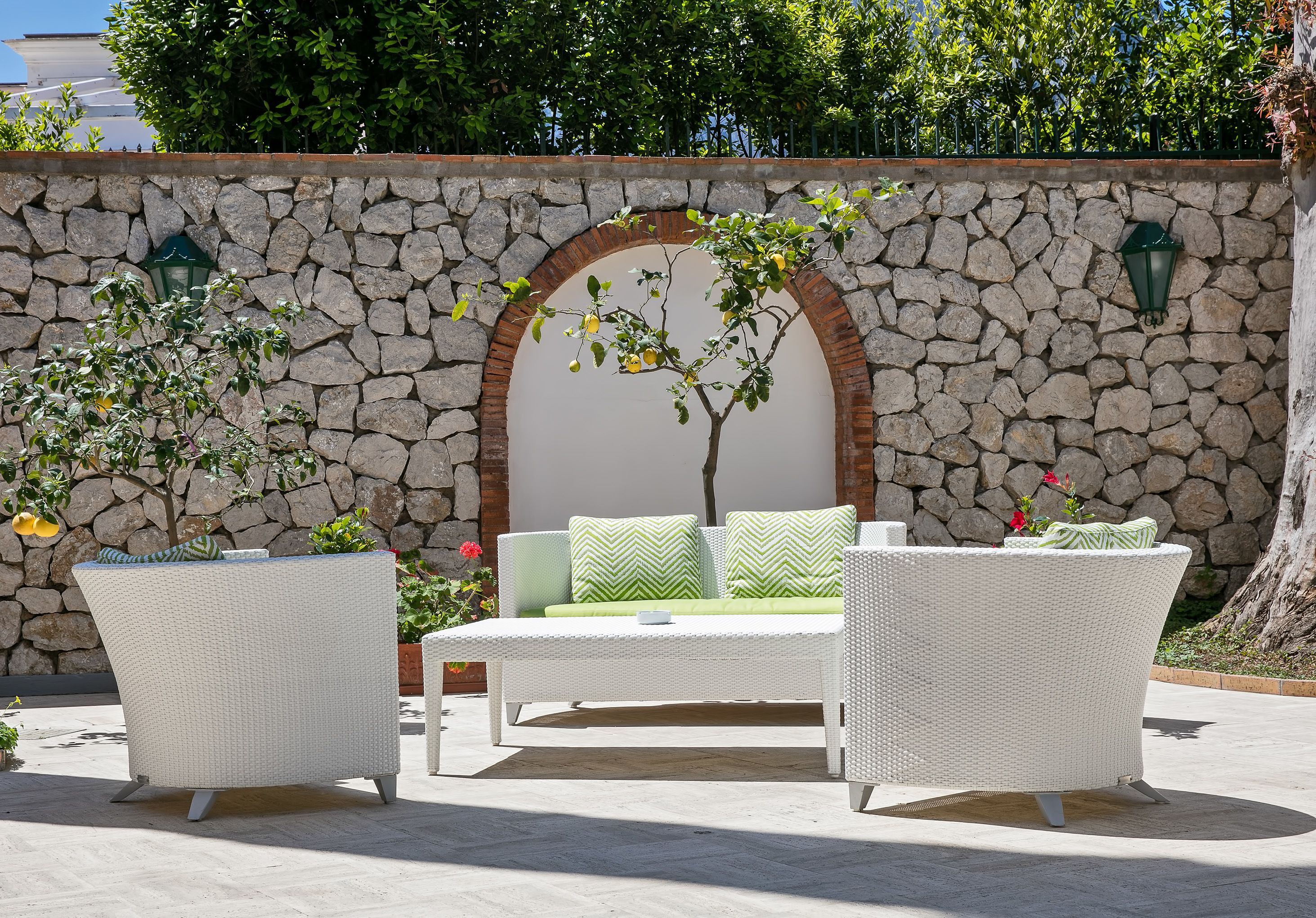 An outdoor seating area with a stone wall, a tree, and a white wicker sofa with green cushions.