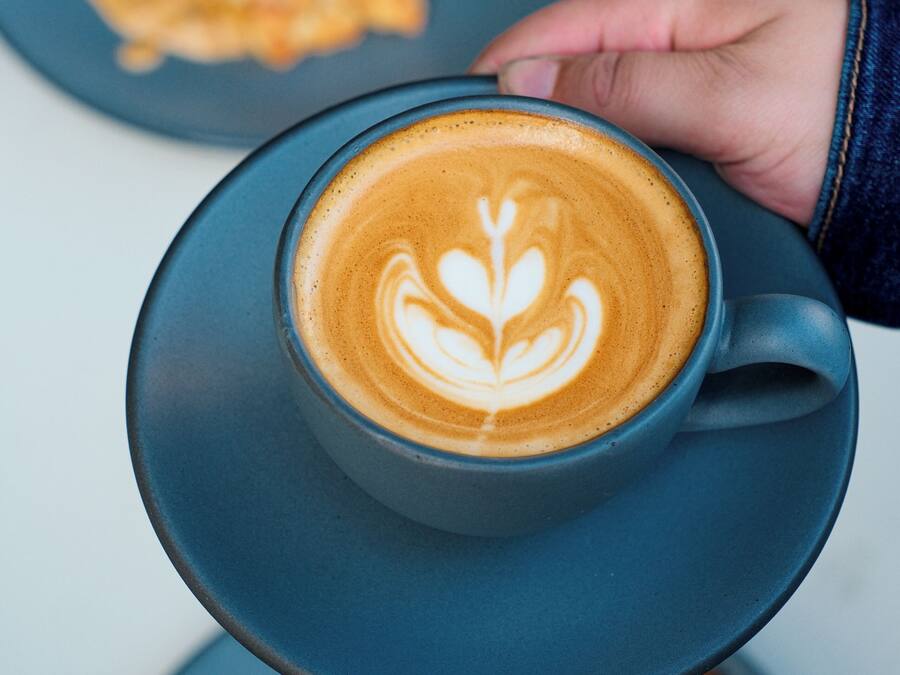 A hand holding a cup of coffee with latte art on a saucer.
