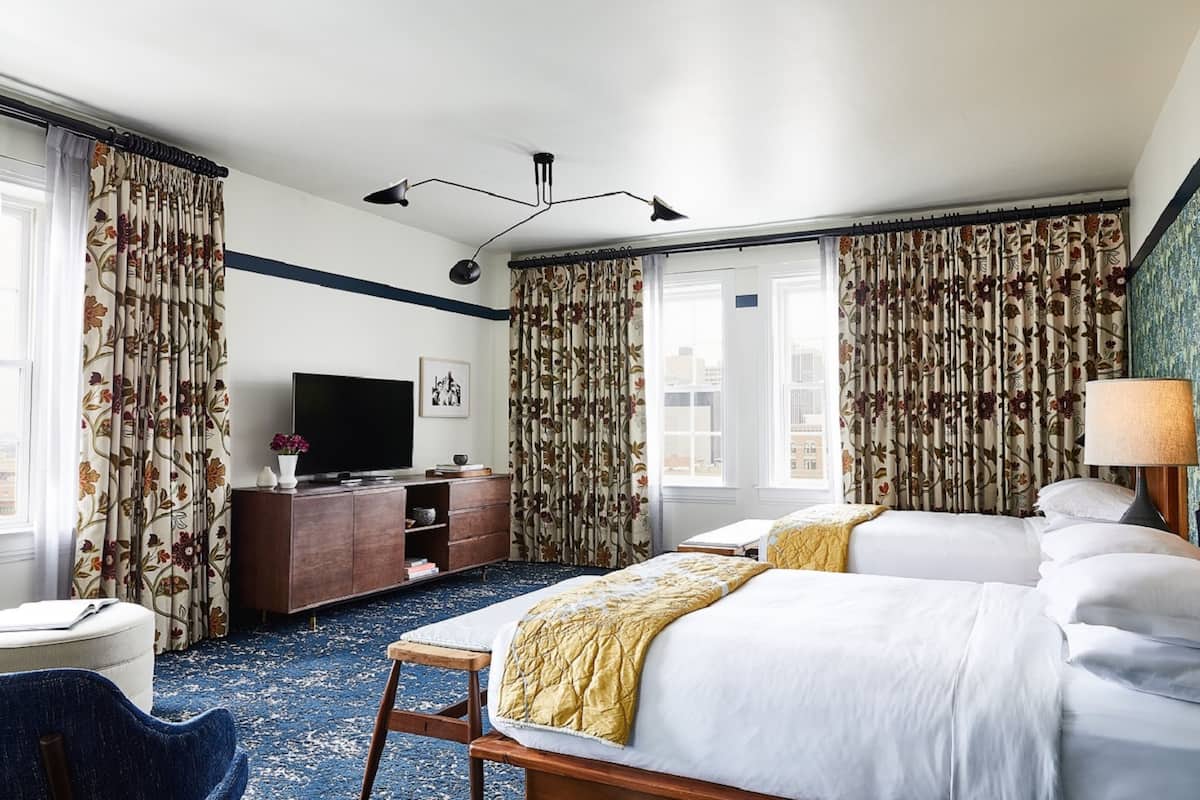 A hotel room with two beds, a desk with a flat-screen TV, a lamp, and a view of the city through large windows.