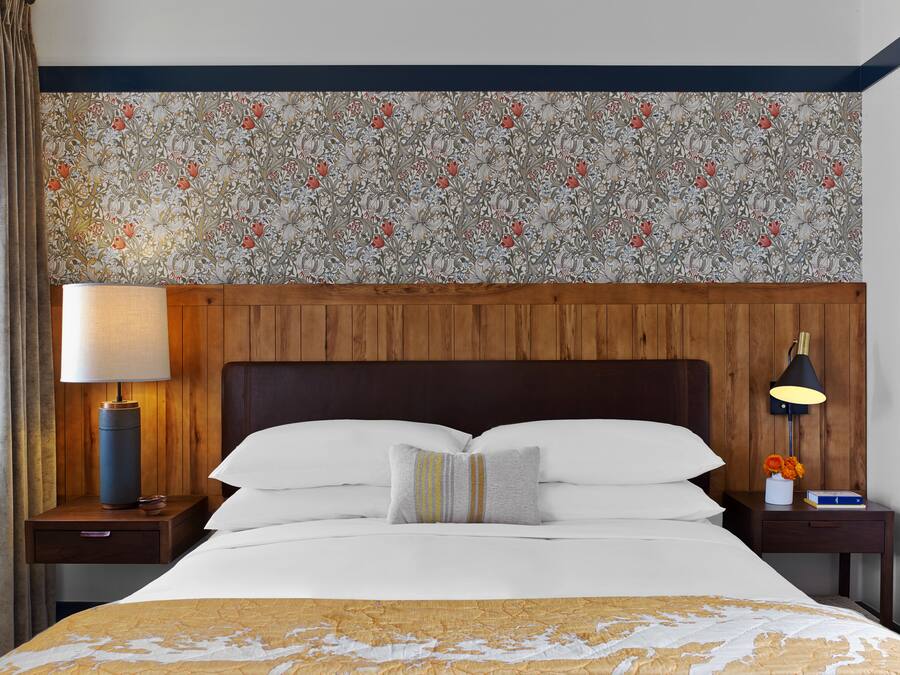 Habitación de hotel con cabecera de madera, una cama grande con ropa blanca, una mesita de noche con lámpara y papel tapiz con estampado floral.