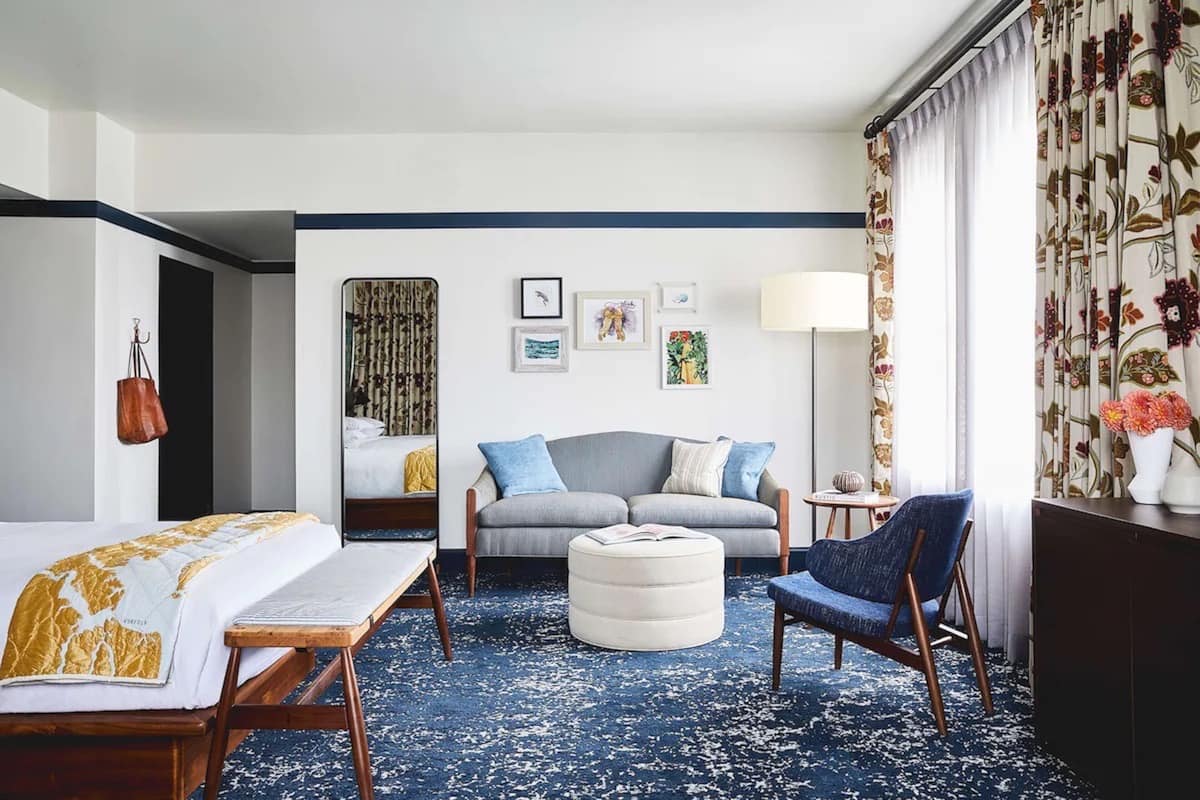 A hotel room with a bed, a sofa, a chair, a round ottoman, a small table, and a lamp. There are wall-mounted pictures and a hanging bag.