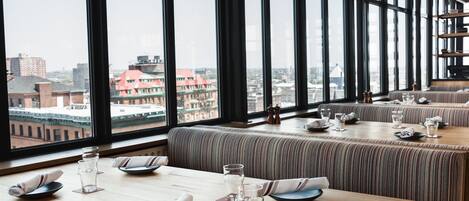 Un restaurante con amplios ventanales que ofrecen vista a la ciudad, mesas de madera con vajilla impecable y asientos con cojines a rayas.