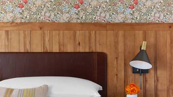 Una habitación de hotel con cabecera de madera, cama con ropa de cama blanca y dorada, mesita de noche con lámpara y un jarrón de flores, y papel tapiz floral.