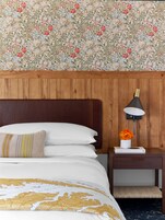 Una habitación de hotel con cabecera de madera, cama con ropa de cama blanca y dorada, mesita de noche con lámpara y un jarrón de flores, y papel tapiz floral.
