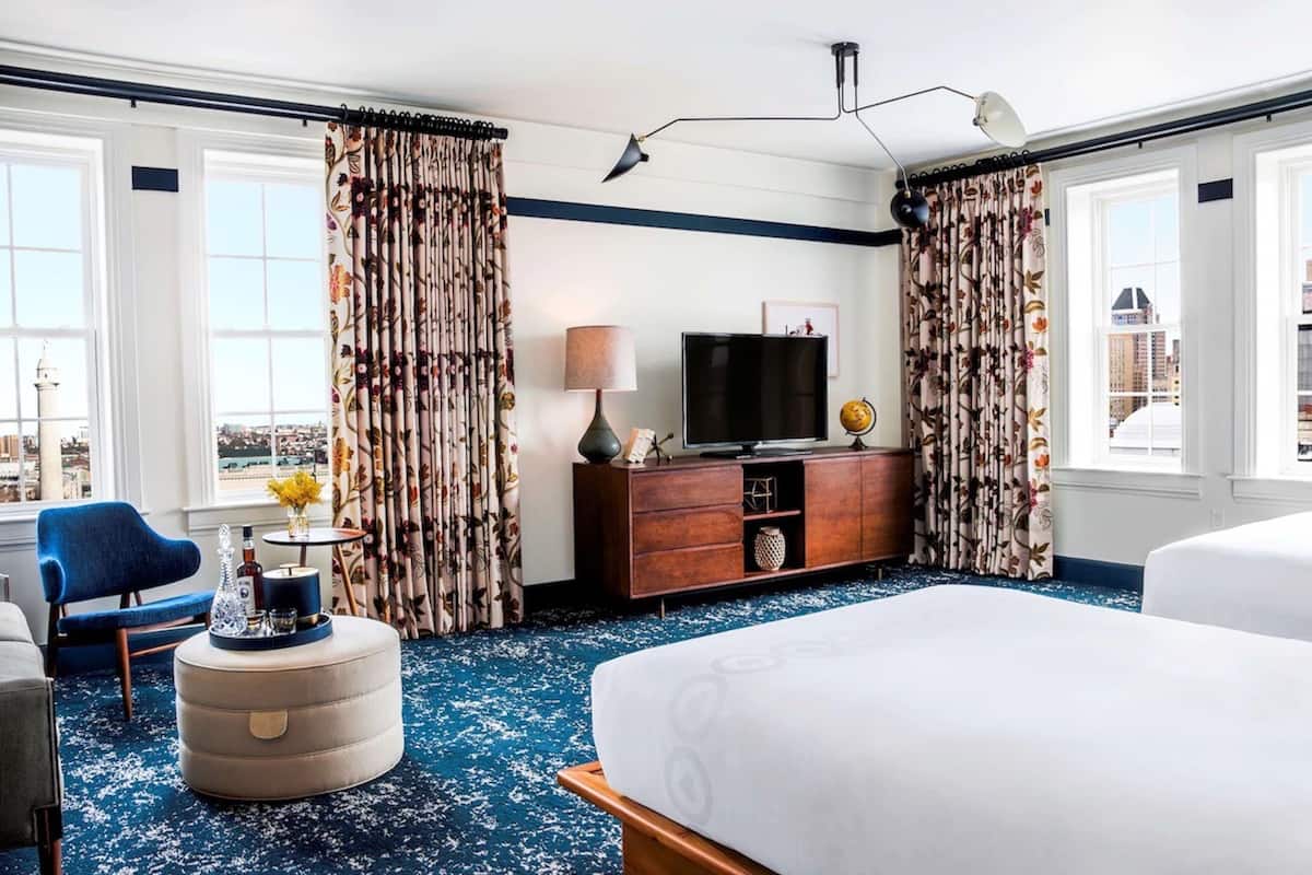 A hotel room with a large bed, a blue armchair, a wooden dresser with a TV, and floral curtains.