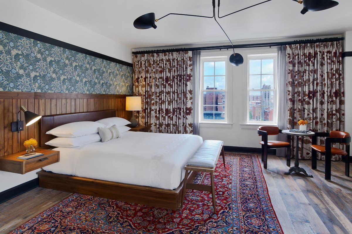 A hotel room with a large bed, two bedside lamps, a small table with chairs, a view of the city through the window, and a patterned carpet.