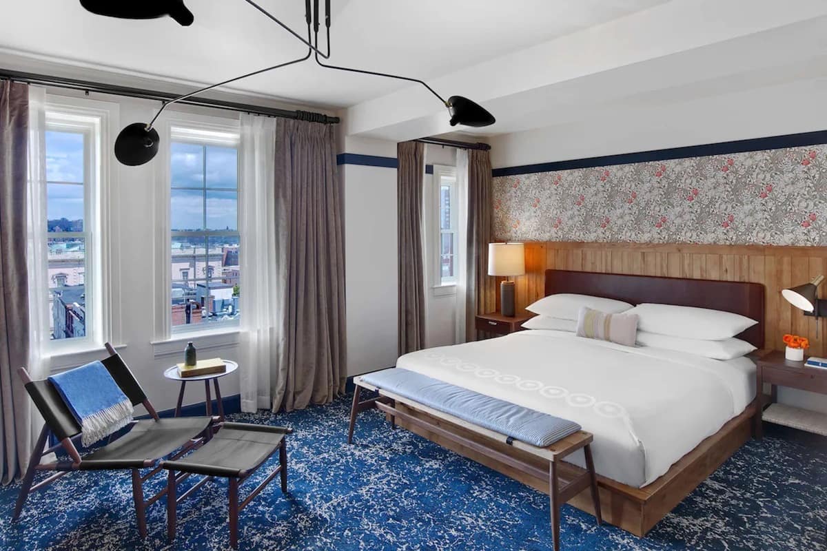 A hotel room with a large bed, a desk, two chairs, a view of the city, and a patterned wallpaper.