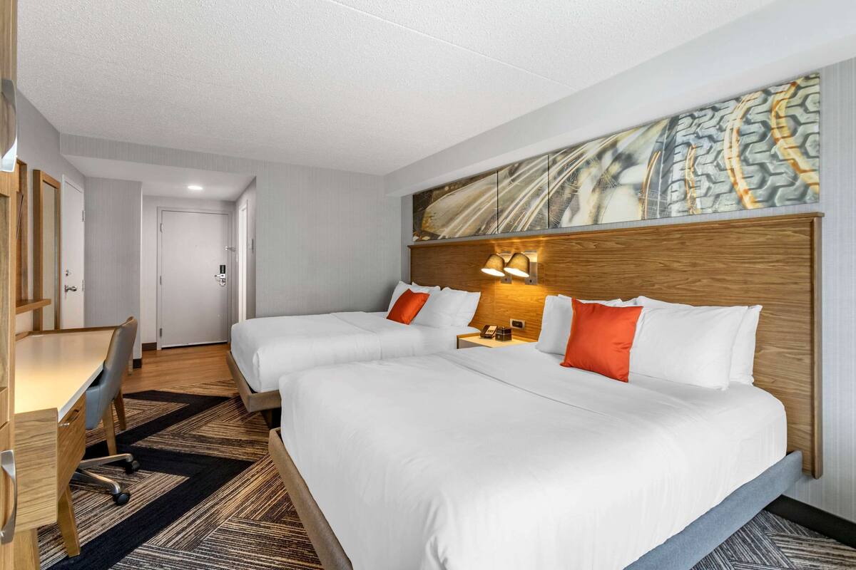 A hotel room with two beds, a desk, and a chair. There is a painting on the wall and a door in the background.