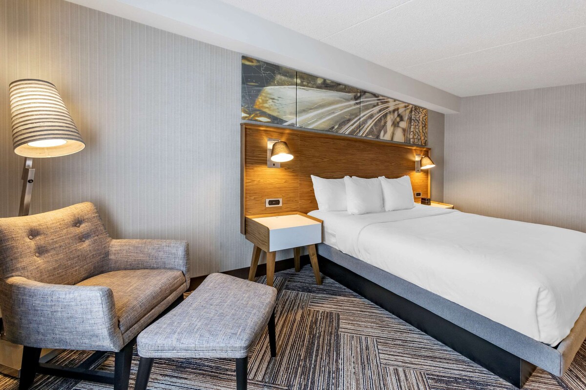 A hotel room with a large bed, a chair, a small table, and a lamp.