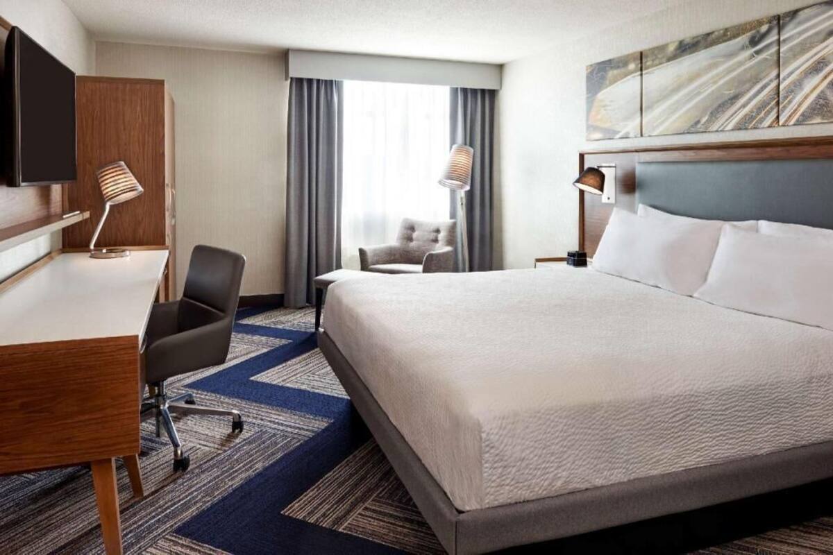 A hotel room with a bed, desk, chair, TV, and artwork on the wall.