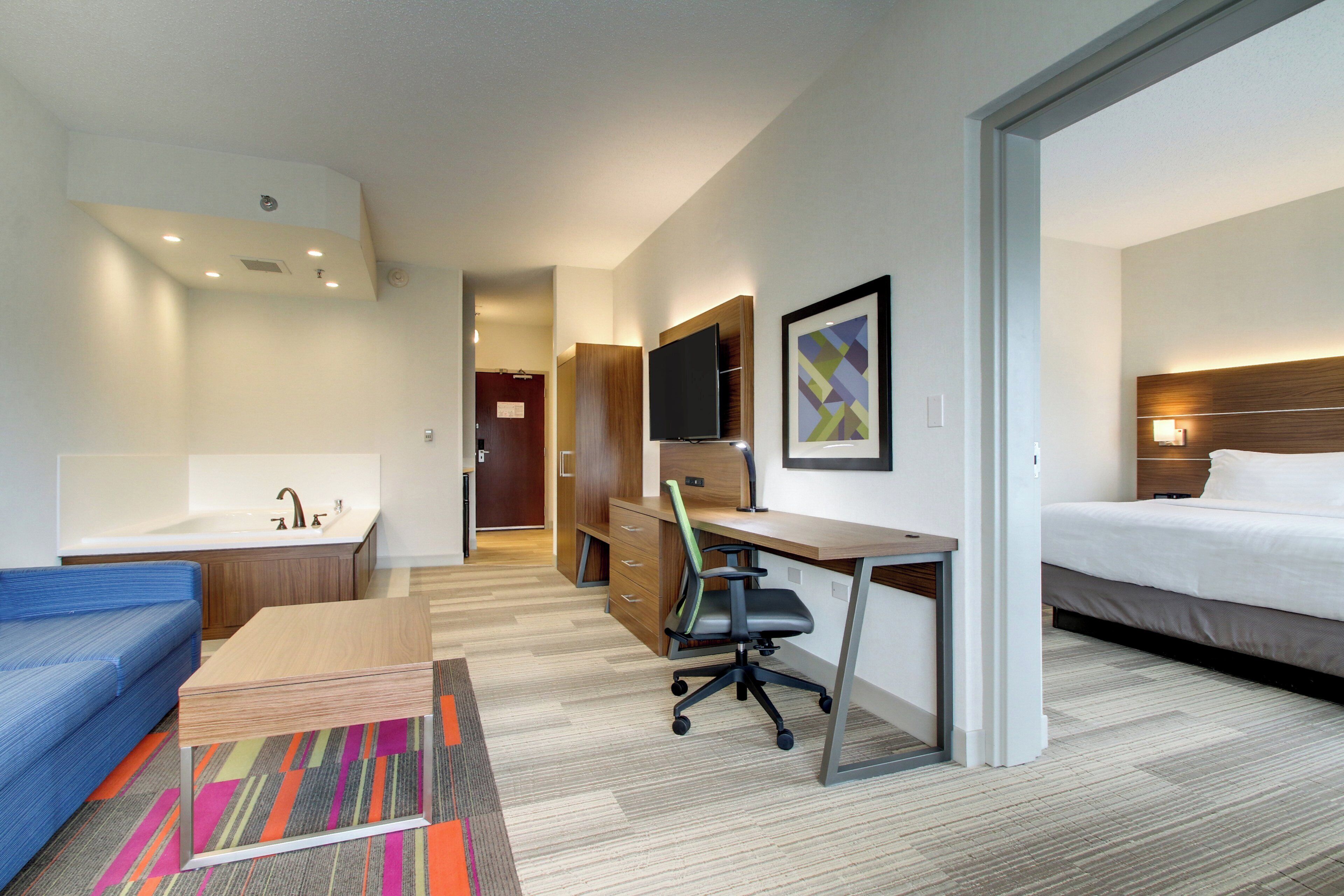 A hotel room with a bed, a desk with a chair, a television, a bathroom with a sink, and a colorful rug.