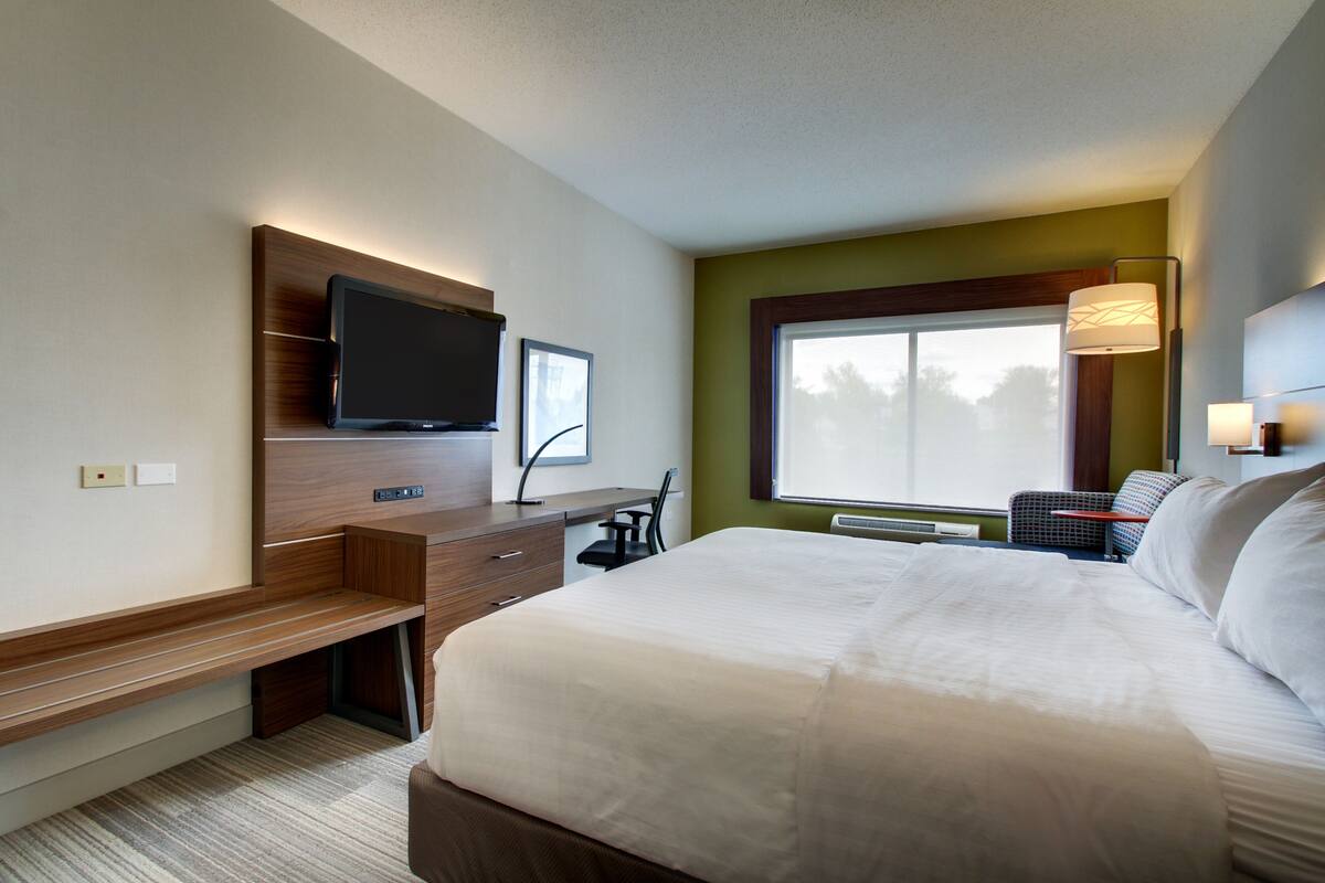 A hotel room with a large bed, a desk with a flat-screen TV, a chair, a window with a view, and a wall-mounted lamp.