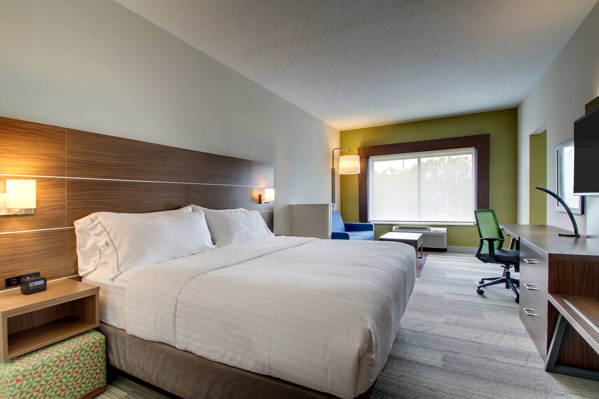 A hotel room with a large bed, bedside table, lamp, desk, chair, and TV.