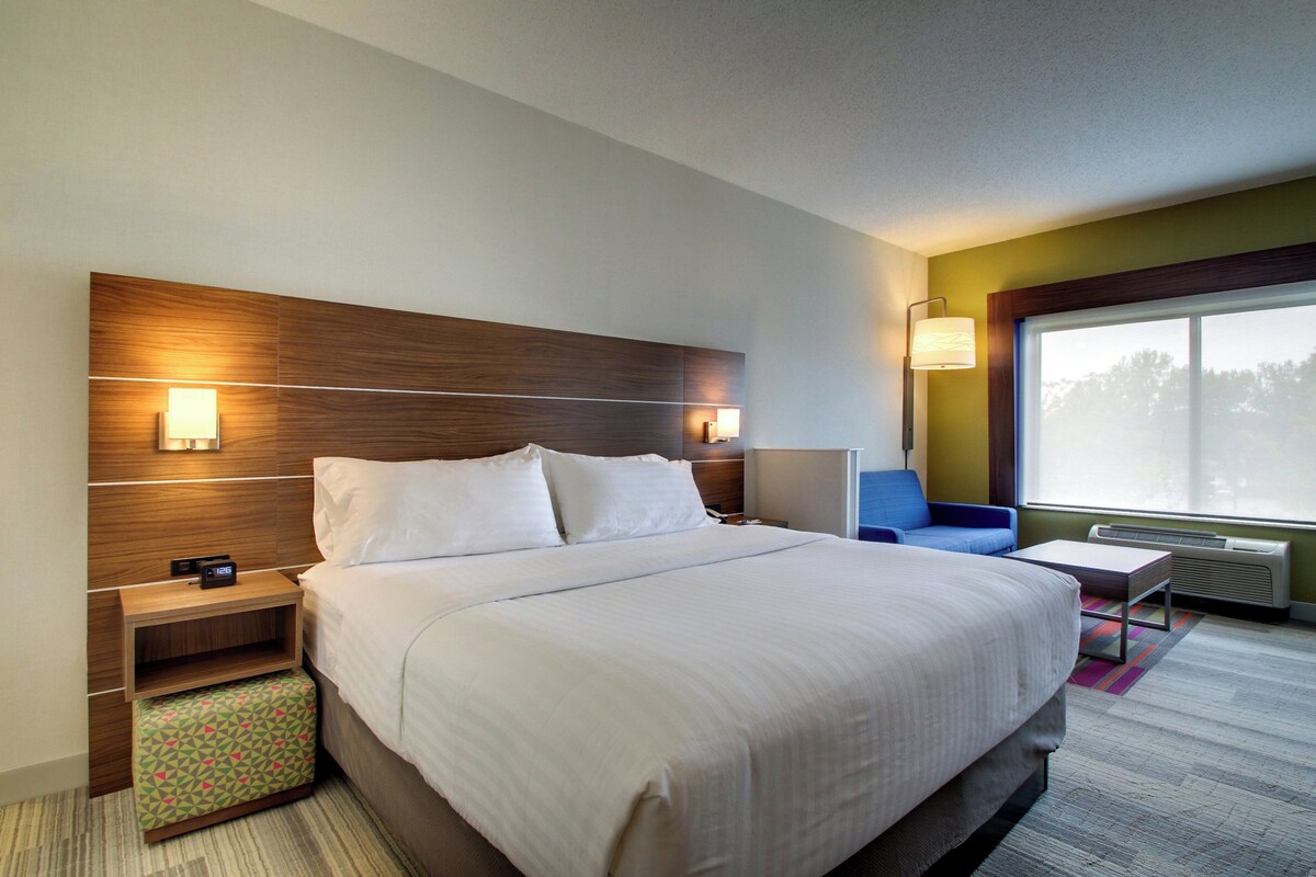 A hotel room with a large bed, bedside tables, a nightstand, a chair, a small table, and a window with a view.