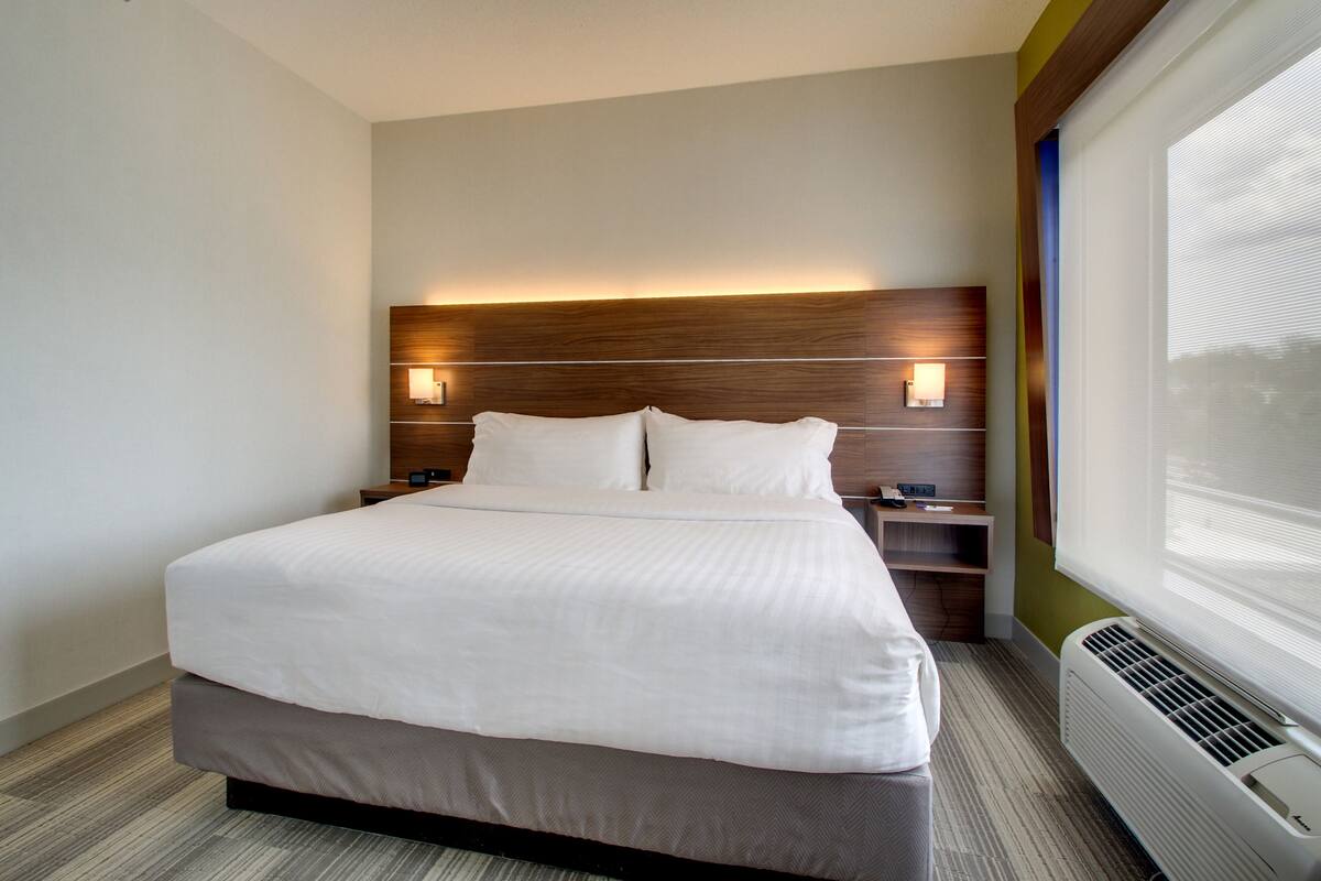 A modern hotel room with a large bed, wooden headboard, bedside tables, and a window with blinds.